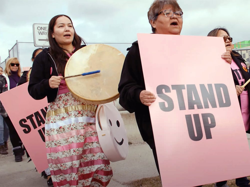 A group of people hold pink signs reading, ‘Stand Up.’ An Indigenous woman wearing a ribbon skirt plays a drum. 