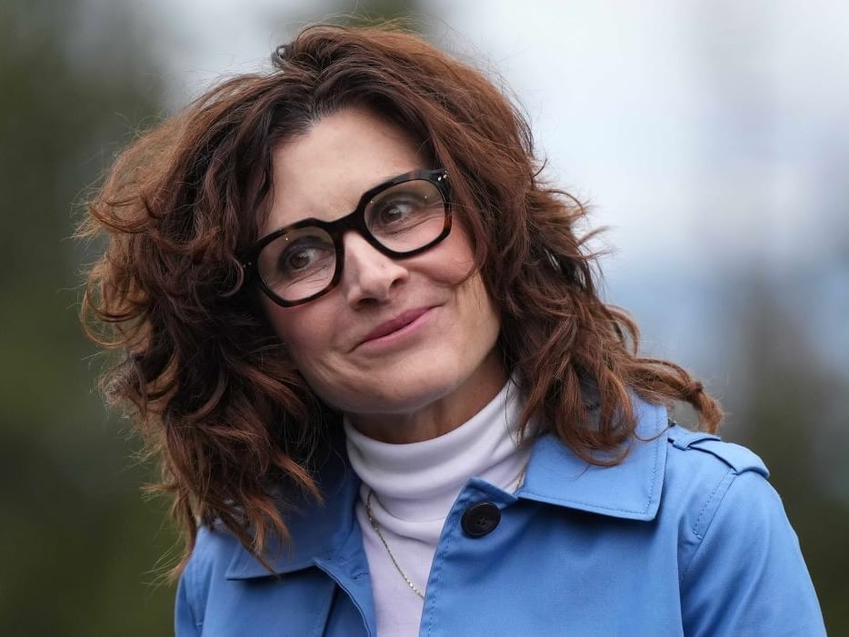 Dallas Brodie, a 50-ish light-skinned woman with tousled brown hair, smiles. She is wearing dark-rimmed glasses, a white turtleneck and pale blue jacket.