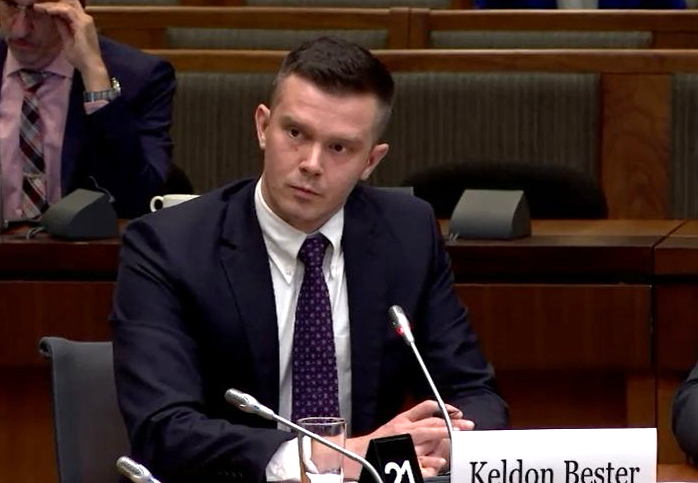 A man with light skin and short brown hair in a dark suit and tie sits in front of a microphone and a sign that reads, ‘Keldon Bester.’