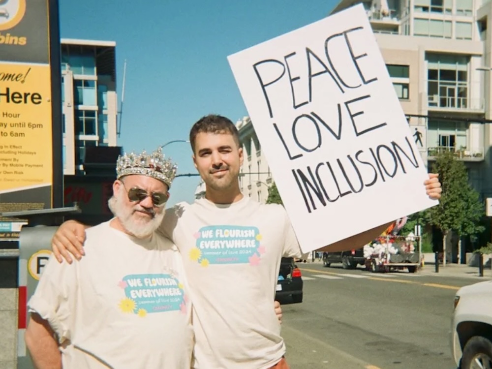A light-skinned man in his 30s with short brown hair and a moustache stands beside a light-skinned man in his 50s who is wearing a crown. The man in his 30s holds up a sign that says ‘Peace, Love, Inclusion.’