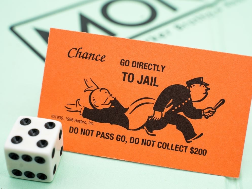 A dice and monopoly playing board with, in the foreground, a ‘Go directly to jail’ monopoly card showing a rich man being carted off by police.