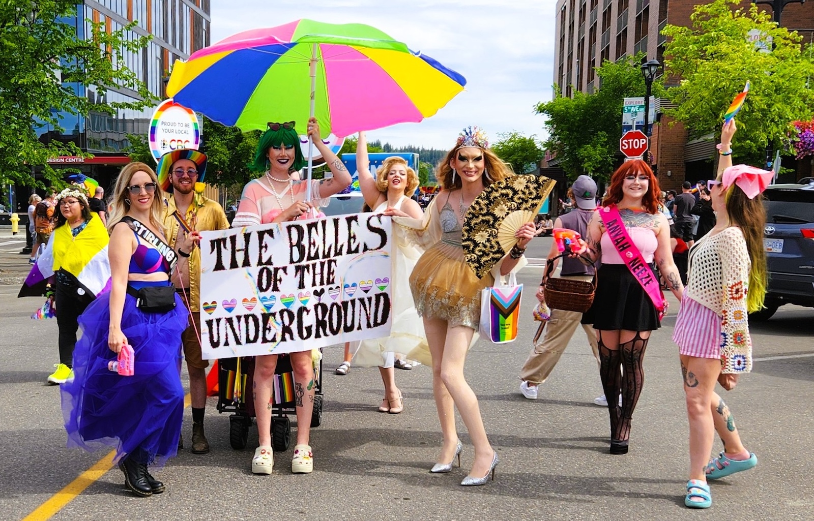 People dressed in colourful skirts and dresses, some with flowing hair and wearing crowns, hold a sign that says ‘Belles of the Underground.’ One person holds a colourful parasol.