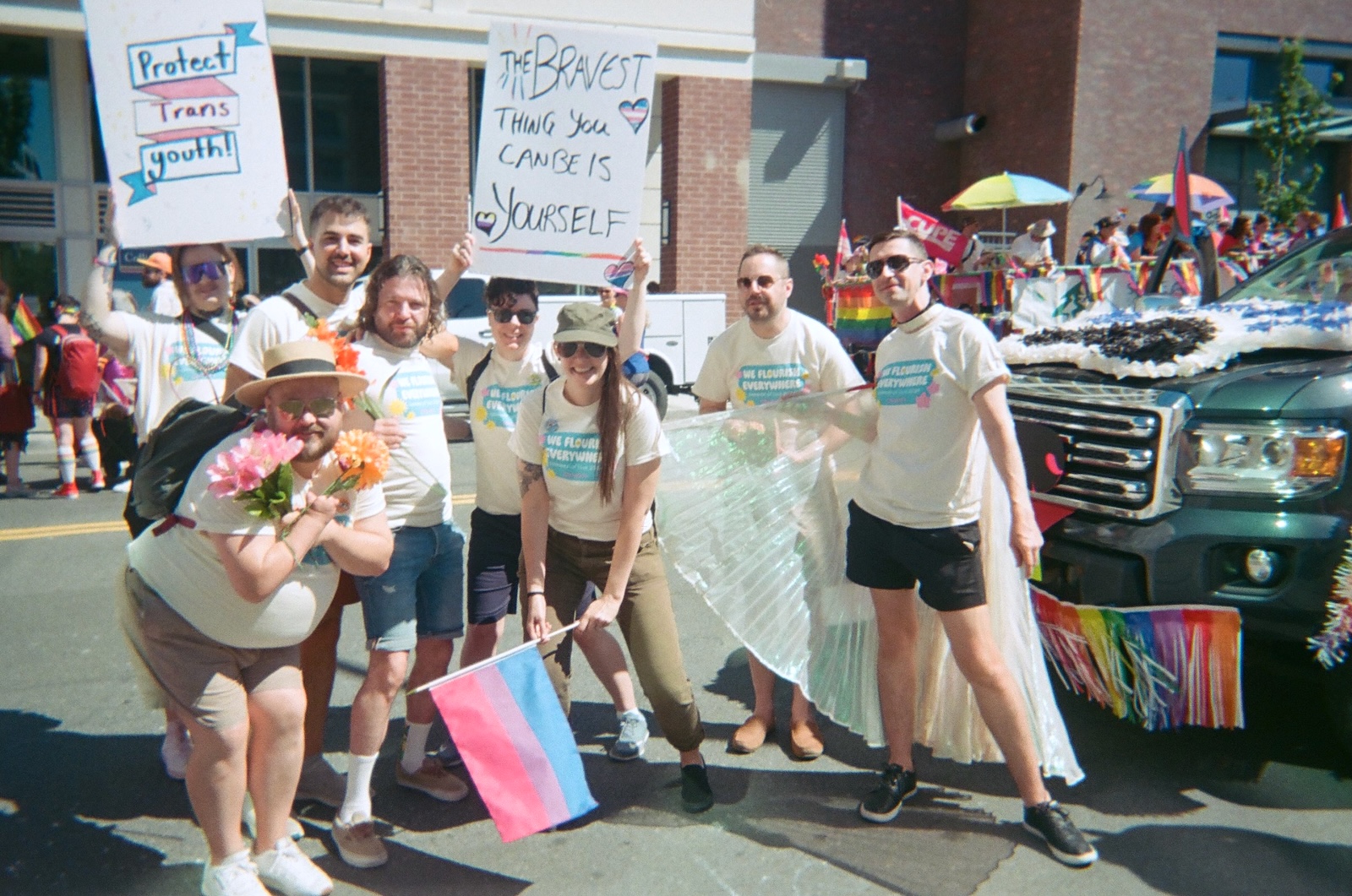 A group of people in white T-shirts pose for a photo. Some hold signs that say ‘The bravest thing you can be is yourself’ and ‘Protect trans youth.’ In the background a decorated truck and rainbow flags can be seen.