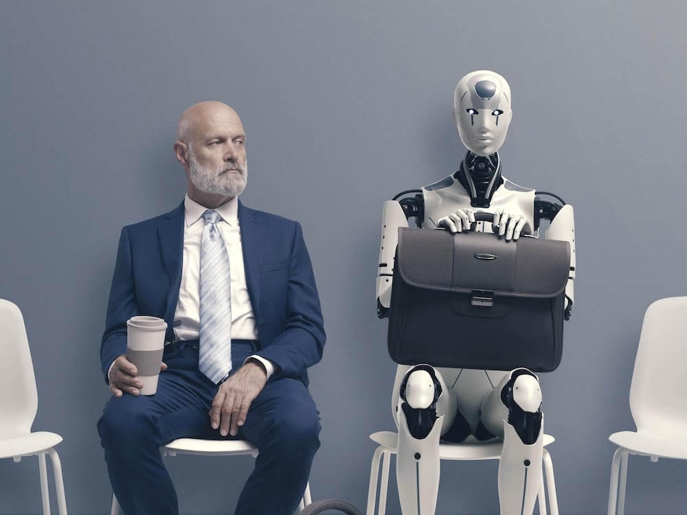 A middle-aged man in a suit sits beside a robot with a briefcase in its lap.