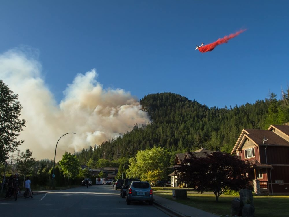 In the foreground, a residential street with large houses and parked cars. In the background, a helicopter drops red fire retardant on a forest fire that has started on a forested hillside, sending a thick plume of grey smoke into the air.