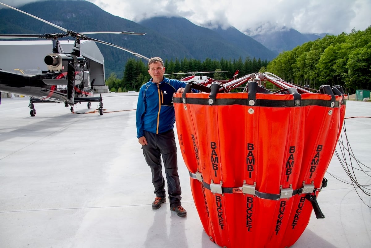 A man with short, light brown hair wearing a blue rain jacket stands beside a very large red bucket that is nearly as tall as he is. A grey helicopter parked on a cement pad can be seen in the background, along with overlapping blue mountains in the far background.