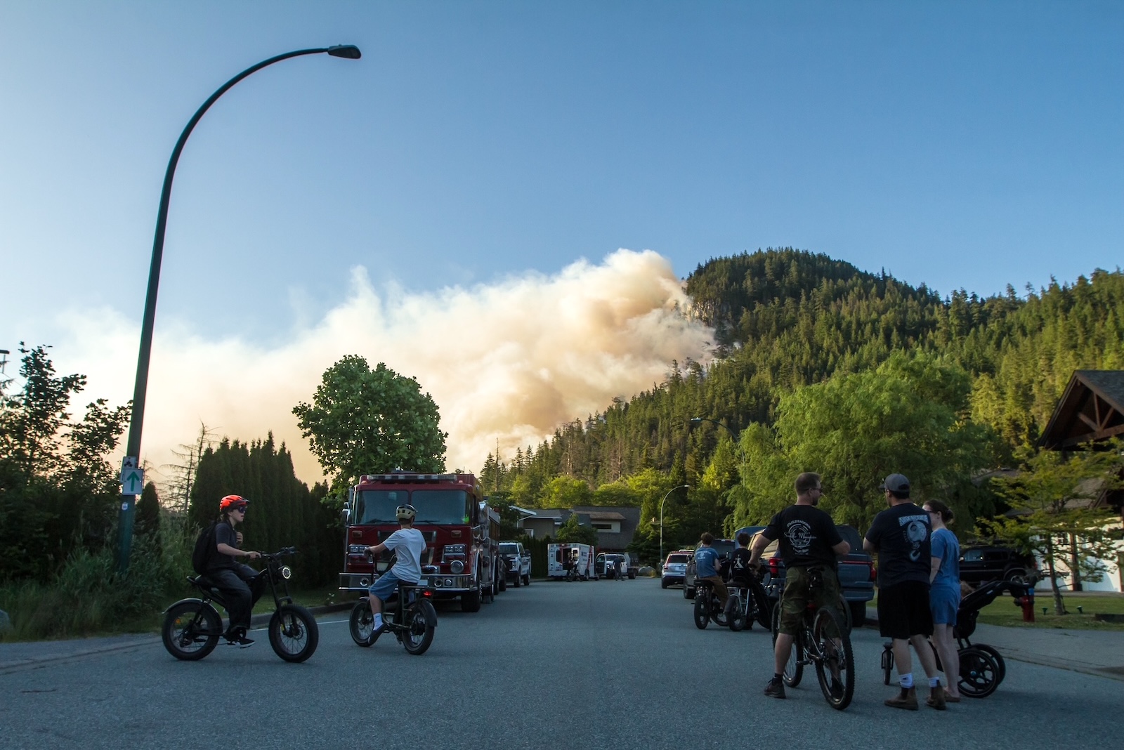 A group of children and adults on bicycles on a suburban street watch a column of smoke on the side of the mountain.