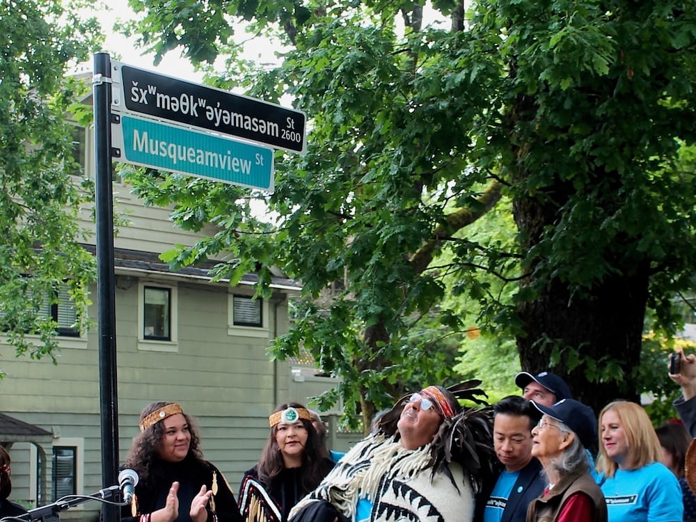 A man wearing glasses and Musqueam regalia looks up at a street sign that says ‘šxʷməθkʷəy̓əmasəm Street.’ To the left of the frame, two women wearing cedar headbands and black clothing decorated with wooden paddles applaud. To the right, more people are gathered, most looking up at the street sign with happy expressions on their faces.