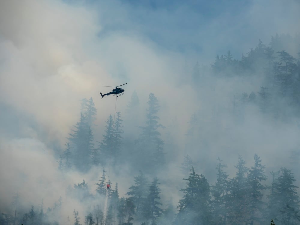 A black helicopter hoisting a large bucket flies over steep slopes shrouded in smoke.