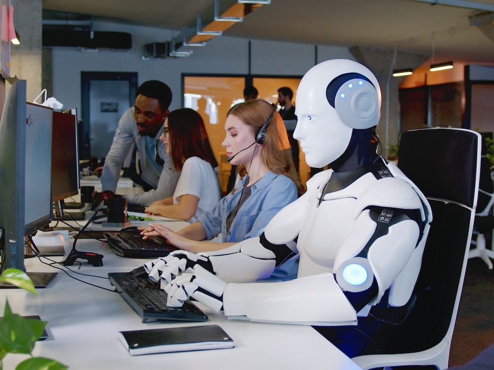 Workers sit in front of computers in an office. Beside them a white robot also sits at a desk typing on a keyboard.