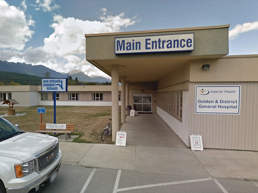 A sign says, ‘Main Entrance.’ Another reads, ‘Golden and District General Hospital.’ The building is one storey and beige, with a mountain in the background.