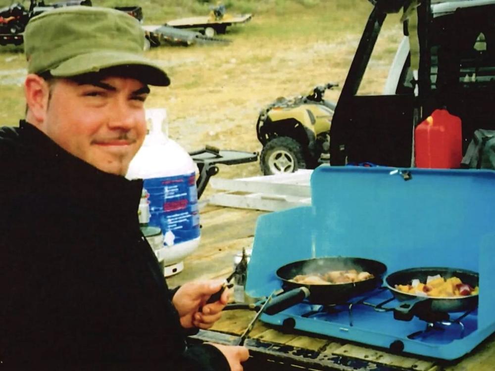 An older, slightly fuzzy photo shows Mazin Zaim cooking vegetables on a camp stove on a pickup truck bed. Zaim has medium-light skin tone and a bit of dark facial hair and is wearing a green cap.