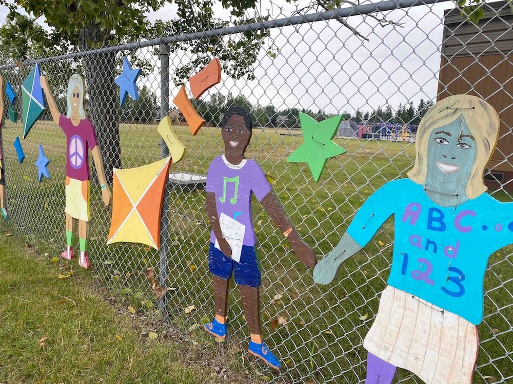 Colourful painted wooden cutouts of children and stars and kites are mounted on a chain-link fence around a Calgary elementary school on a grey day.