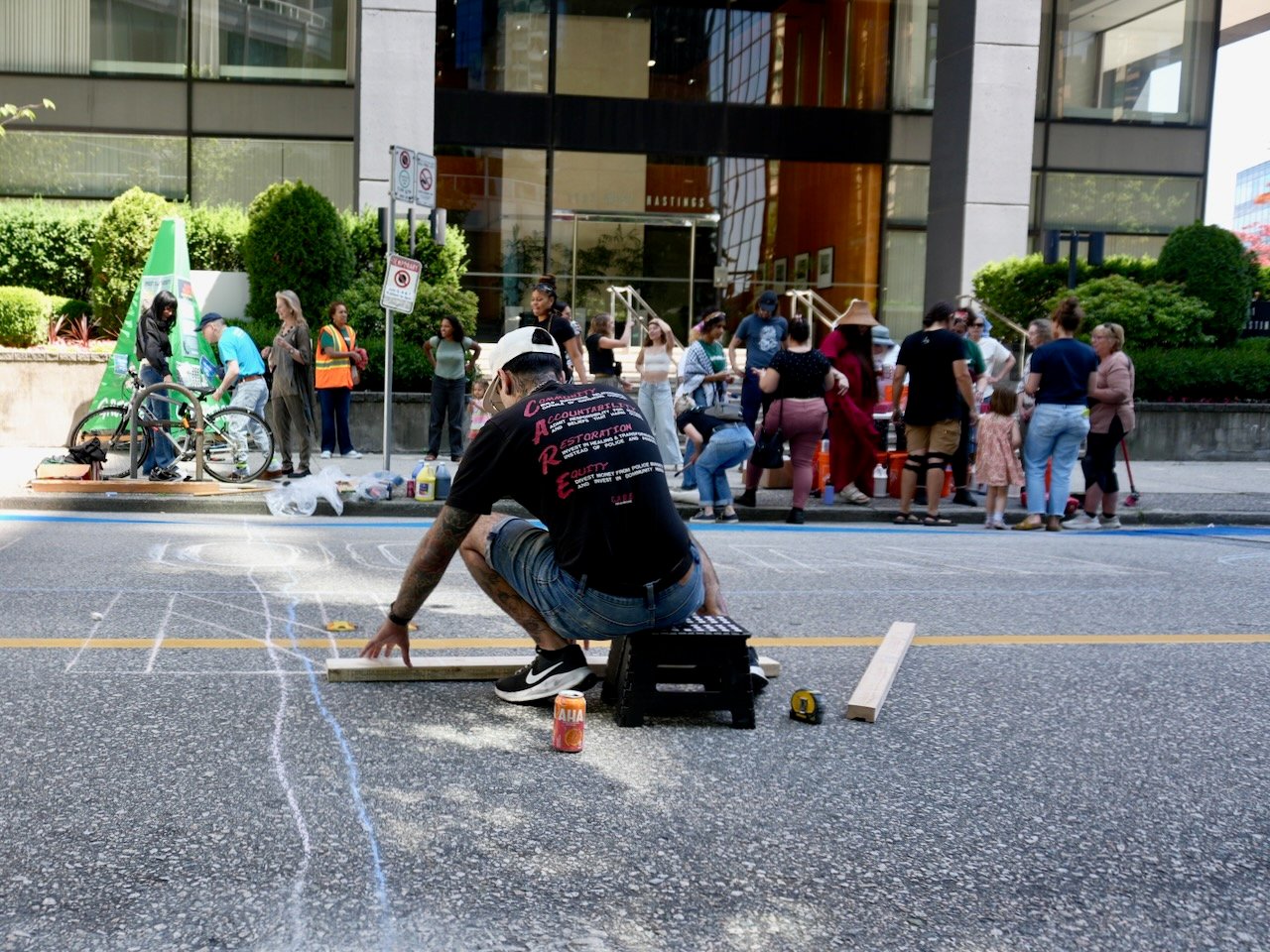A man in a black baseball cap and a black t-shirt crouches over a sidewalk, measuring and using chalk to draw. In the background a group of people can be seen.