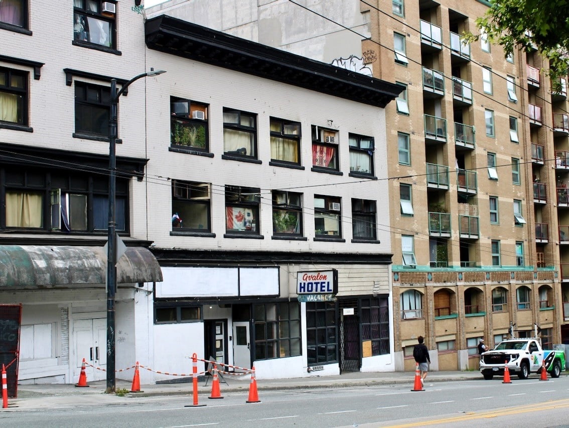A street in downtown Vancouver. In the middle of the photo, between two taller buildings, is a white three-storey building with a sign that says ‘Avalon Hotel.’