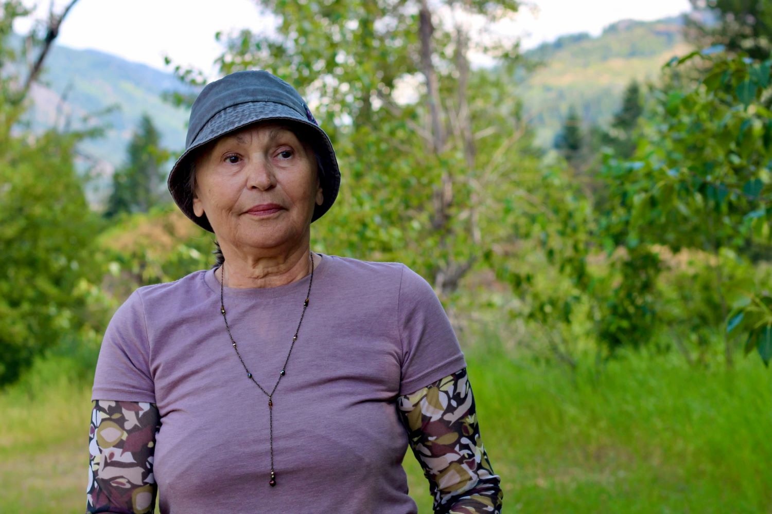 A 50-ish woman with medium-light skin tone stands outside. She wears a blue bucket hat and a lilac-coloured shirt.