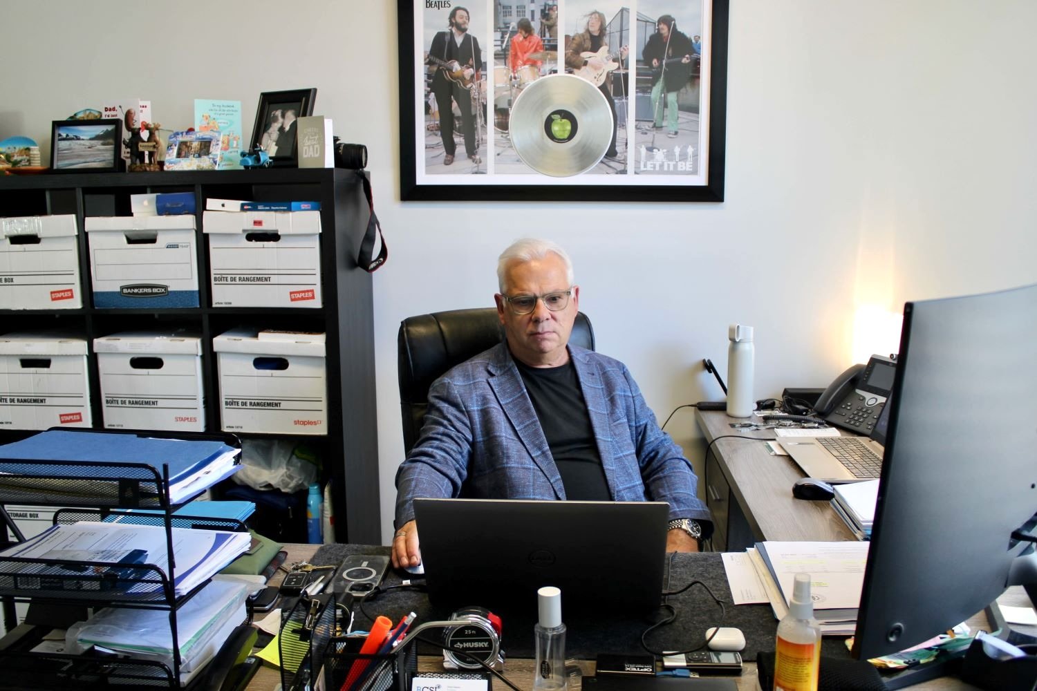 A light-skinned middle-aged man with white hair sits at a desk with boxes of files behind him. He wears a black T-shirt and a blue sports jacket.