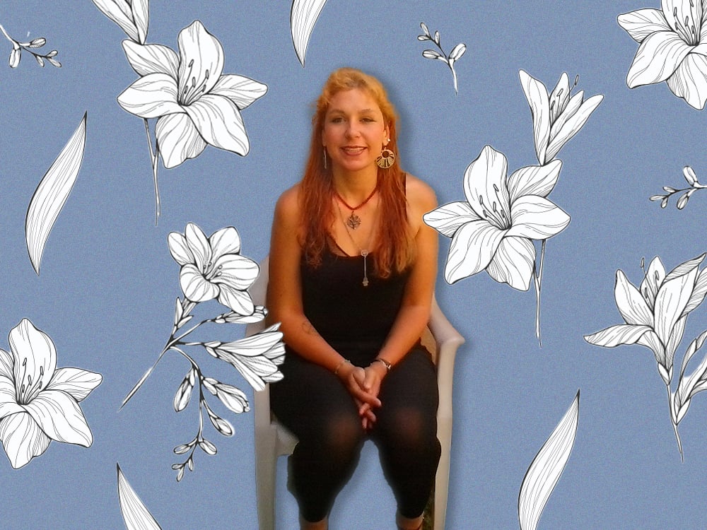 A young woman with long reddish-blond hair smiles at the camera, seated in a white chair. She wears a necklace and a black top and slacks. Her photo is superimposed on a purple background with white flowers.