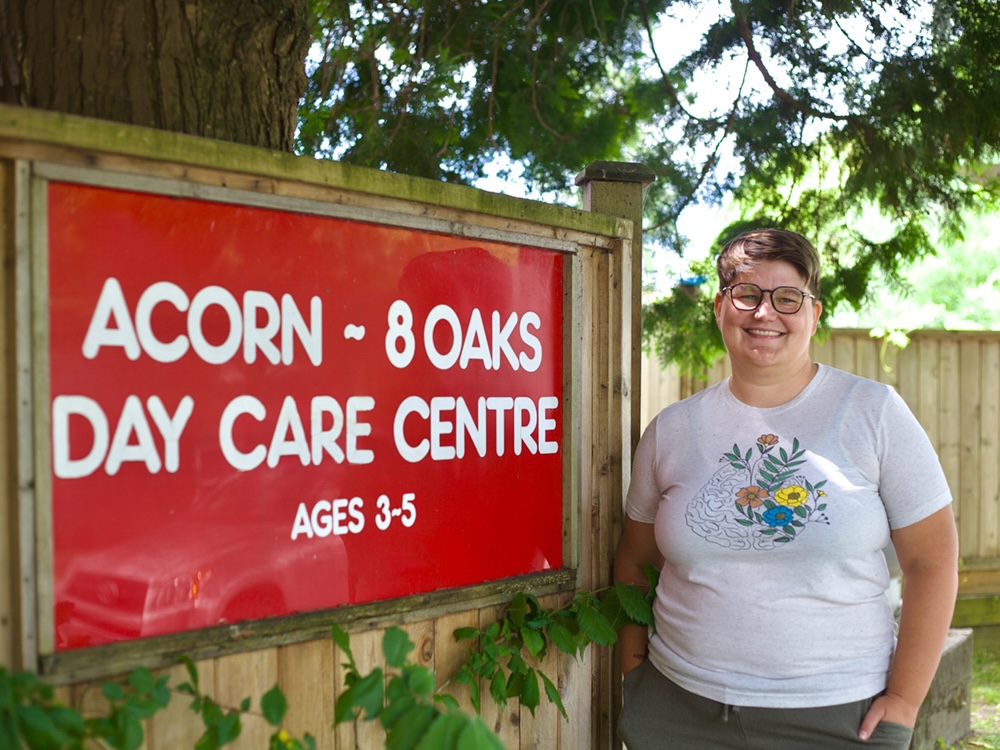 A person with short brown hair and light skin tone wears a T-shirt and glasses and stands in front of a sign that reads, ‘Acorn 8 Oaks Day Care Centre, Ages 3 to 5.’