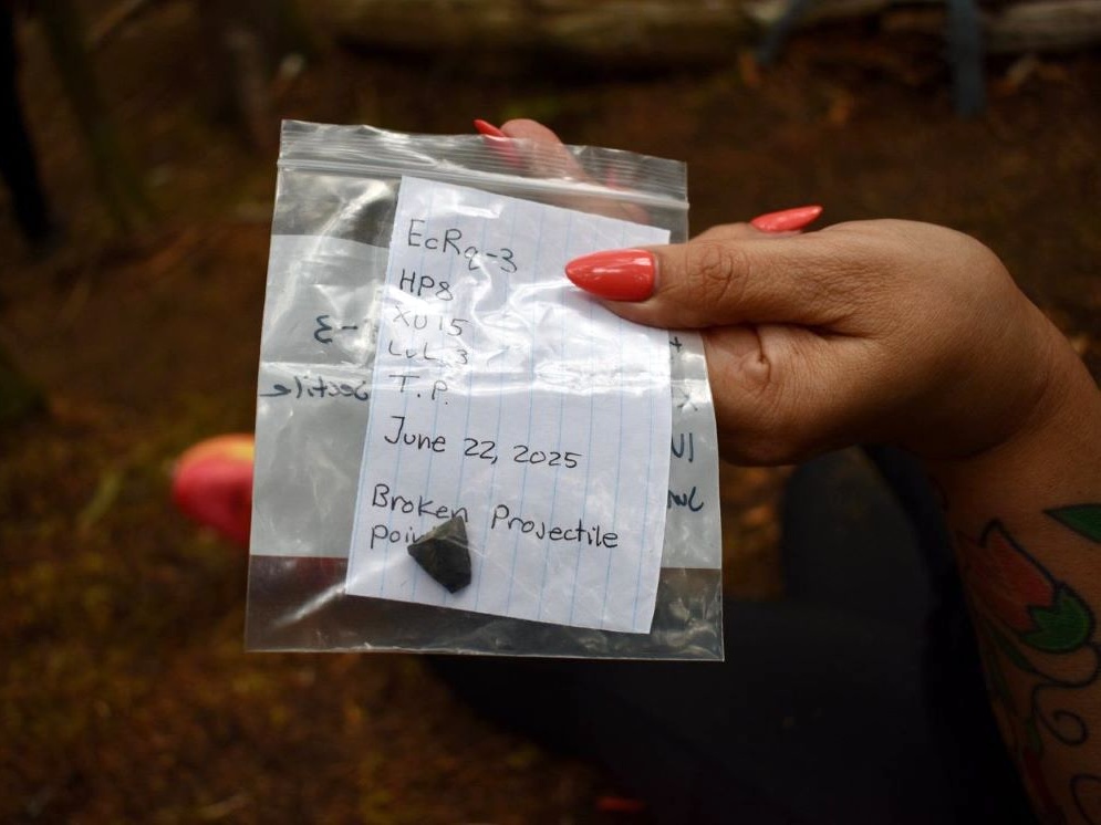 A woman’s hand with long orange nails holds a clear plastic bag that holds an artifact made of rock with notes written on a white piece of paper.