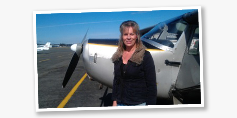 A woman with long blond hair and light skin tone stands in front of a small airplane.