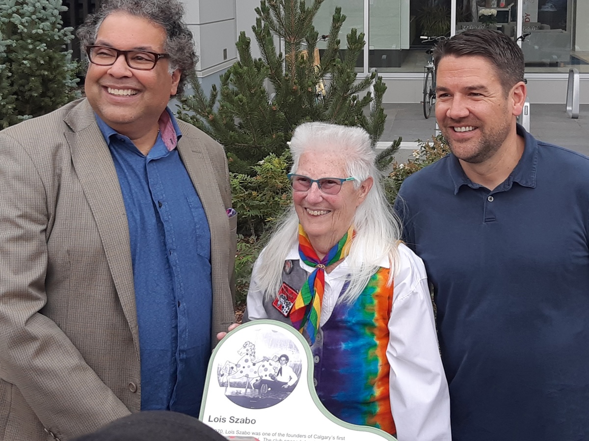 Lois Szabo, an older woman with a silver mullet and light skin tone, stands outside with former Calgary mayor Naheed Nenshi, a man with medium skin tone and dark hair, and former city councillor Evan Woolley, a man with light skin tone and dark hair.
