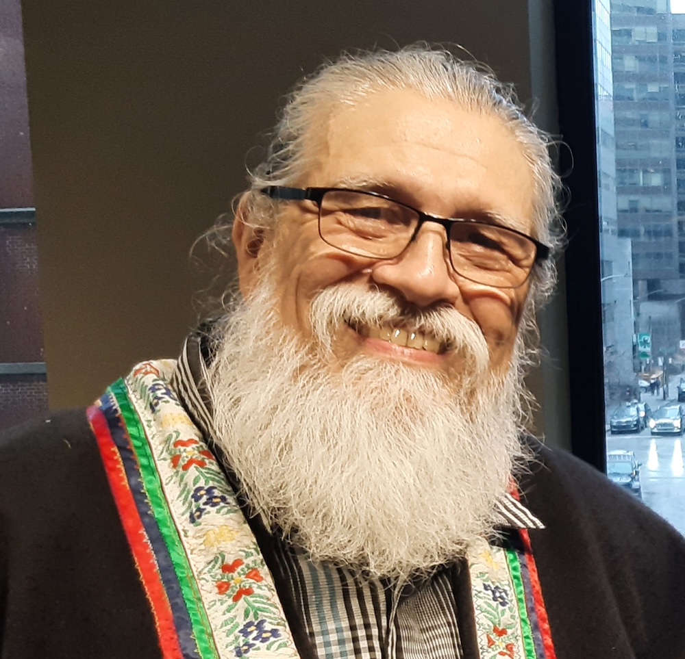 A person with medium-light skin tone and white hair and a beard wears glasses and smiles broadly at the camera.