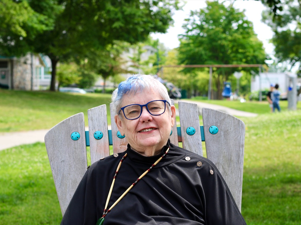 barbara findlay, an older woman with light skin tone and short grey hair that has a blue streak, sits in a chair in a park.