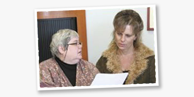 barbara findlay, on the left, an older woman with short grey hair and light skin tone, looks towards Kimberly Nixon, on the right, a younger woman with light skin tone and shoulder-length blond hair. The two are considering information on a sheet of paper.