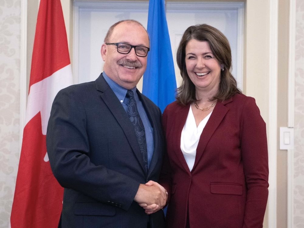 A 66-year-old light-skinned man with a moustache, wearing a suit, smiles as he shakes hands with Danielle Smith, a woman in her mid-50s wearing a white shirt and burgundy blazer.