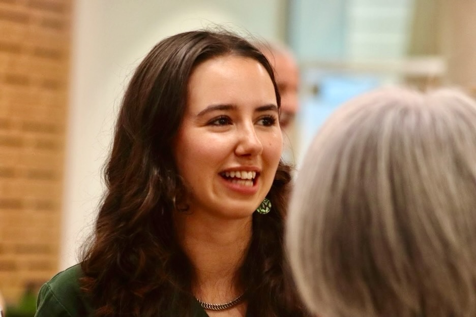 A young woman with long wavy brown hair smiles and speaks to potential supporters at an event.