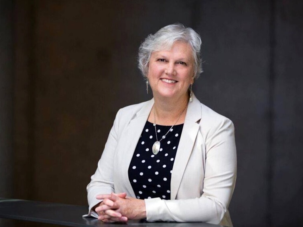 A 50-ish light-skinned woman with short grey hair and an oatmeal-coloured suit smiles at the camera.
