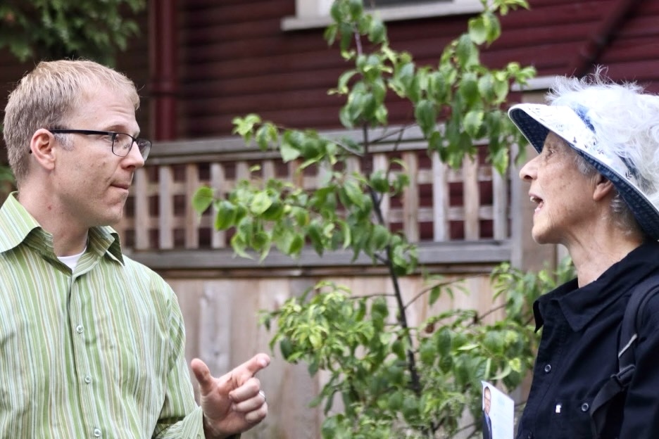 A light-skinned man in his 40s with blond hair and glasses speaks to an older woman wearing a hat in an outdoor setting.
