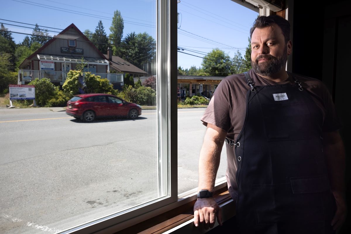 A man with light skin tone and dark brown hair and beard, wearing a black apron over a brown T-shirt, stands by a window looking out on a small-town street.