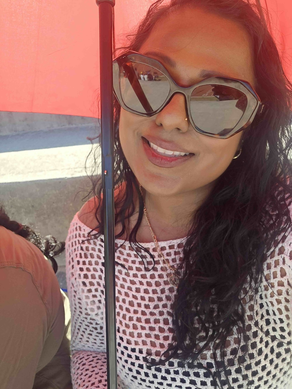 Cheyanne Ratnam has long wavy dark hair and a medium skin tone. She is wearing sunglasses and a white mesh top. She is seated outside under a red umbrella on a bright day.