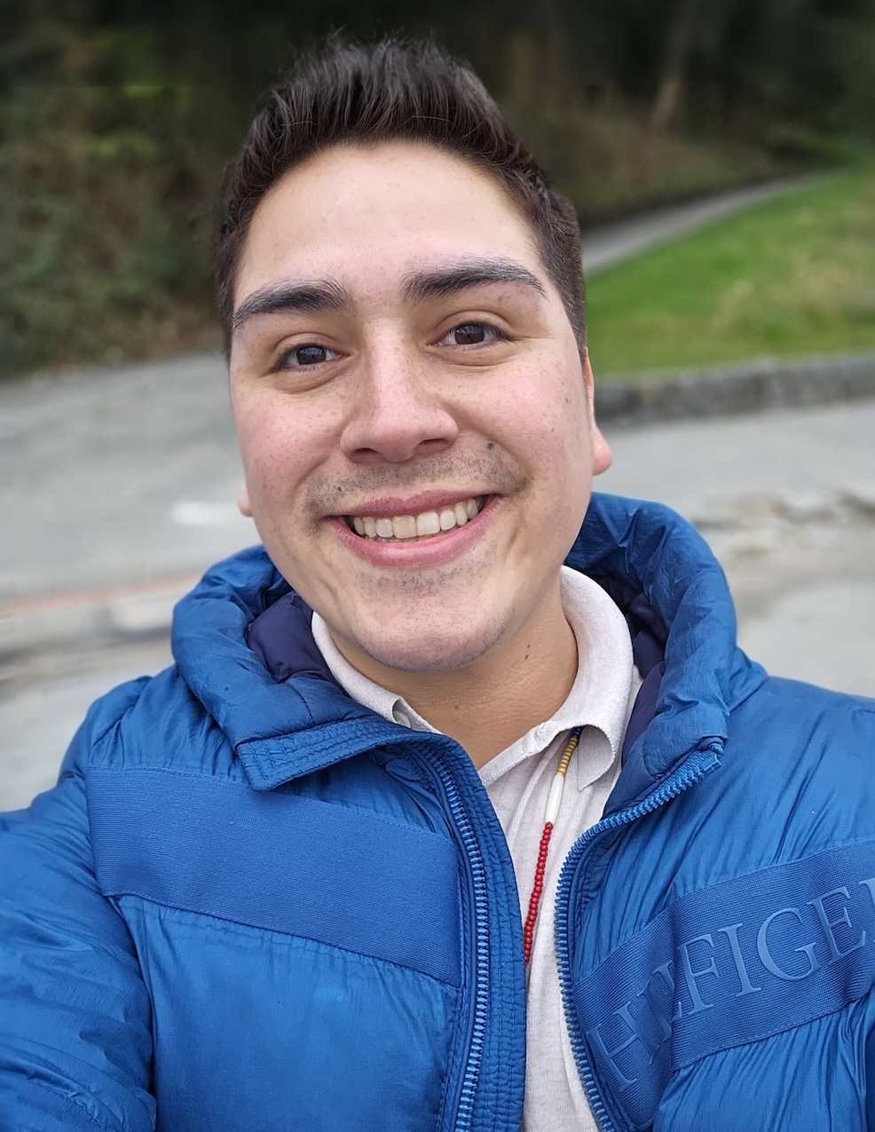 Gage Sabattis has short, spiky dark hair and a light skin tone. He is smiling at the camera and wearing a blue puffer jacket over a light cotton shirt.