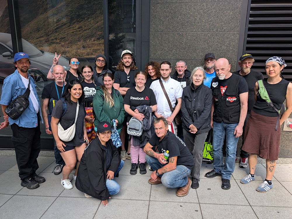 A group of casually dressed people pose for a photo while standing on the sidewalk. Their expressions are cheerful.