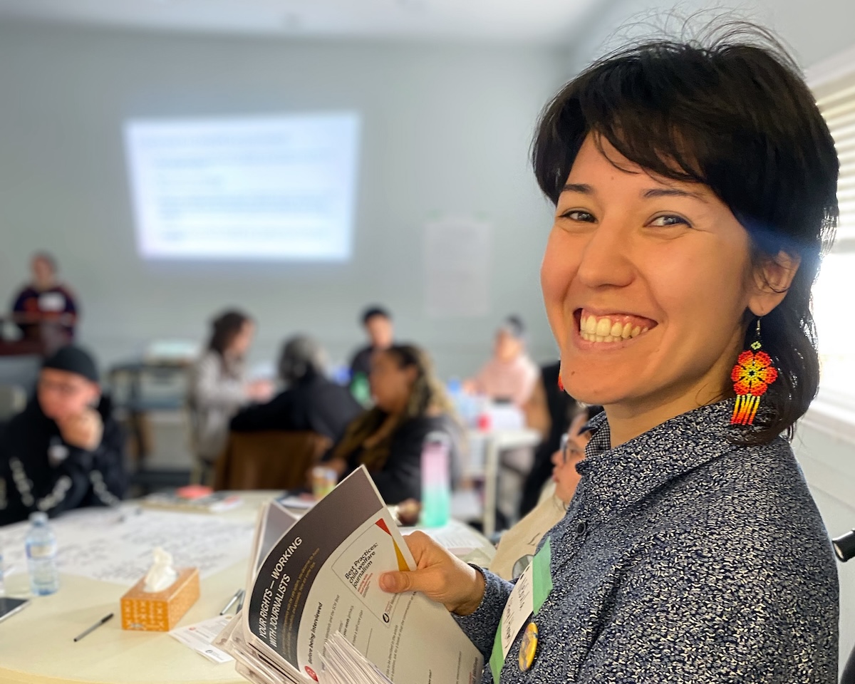 Nebula Shen has shoulder-length dark hair and a medium skin tone. They are turning slightly to smile widely at the camera. They are wearing colourful beaded floral earrings and a grey and white patterned top. They are holding a stack of papers and standing in an indoor space where people are gathered around circular tables.