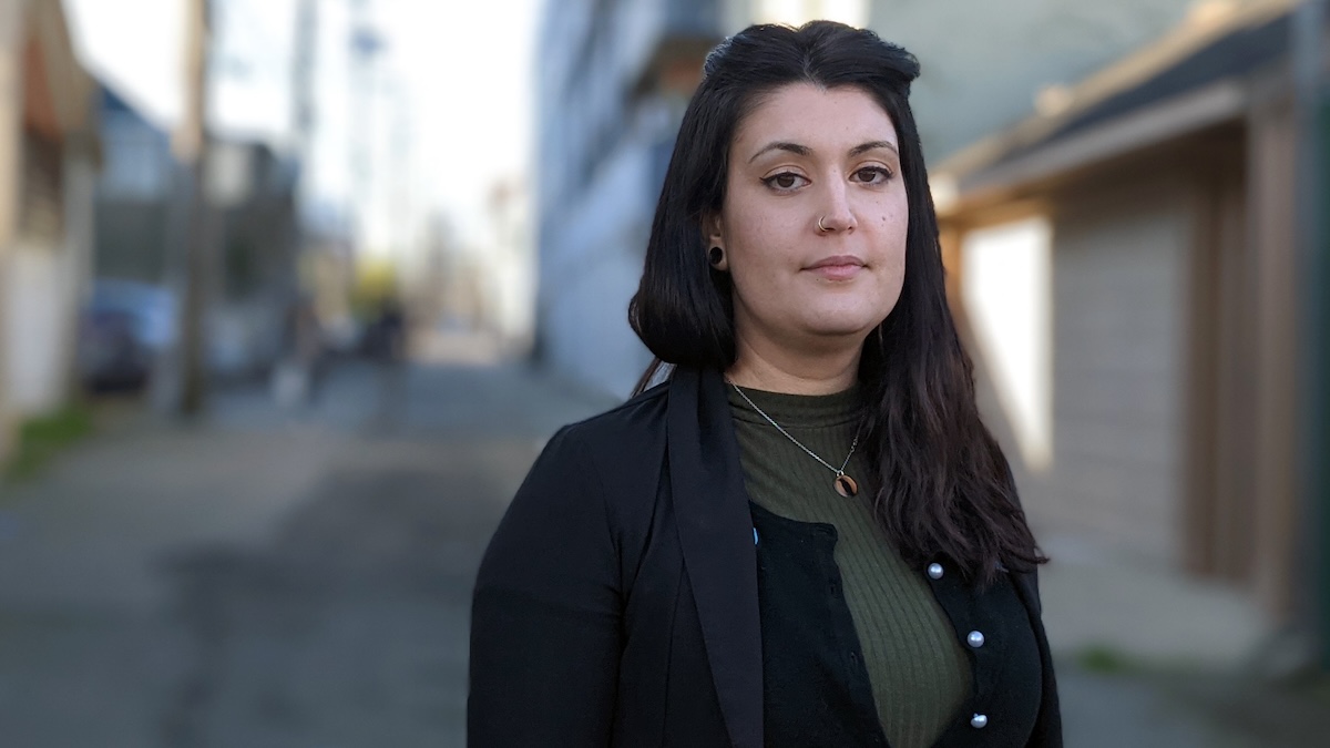Nina Taghaddosi has long dark wavy hair and a pale skin tone. She is standing in an alley and looking directly at the camera. She is wearing a black jacket over an olive-coloured top.