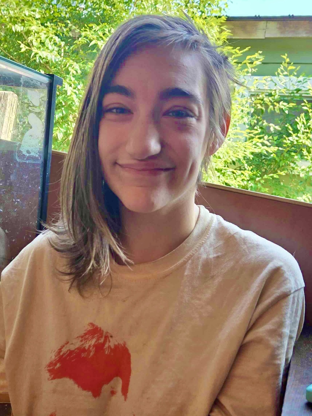 River Ravenswood has shoulder-length light brown hair and a medium skin tone. They are smiling at the camera and seated indoors on a sunny day. They are wearing a beige T-shirt with a graphic of a red mountain on the chest.
