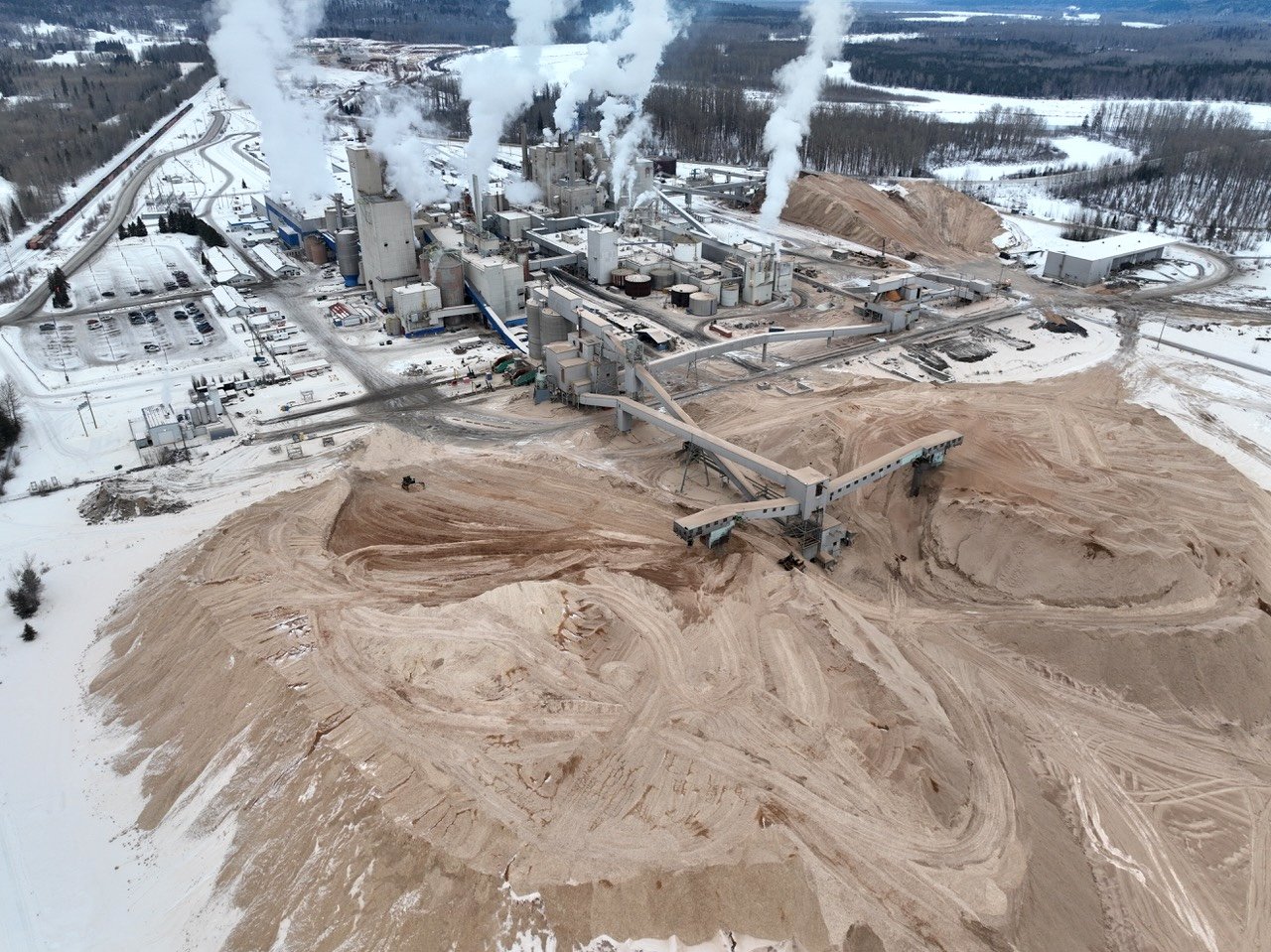 An aerial view of mountains of wood chips in front of and to the right of a pulp mill.