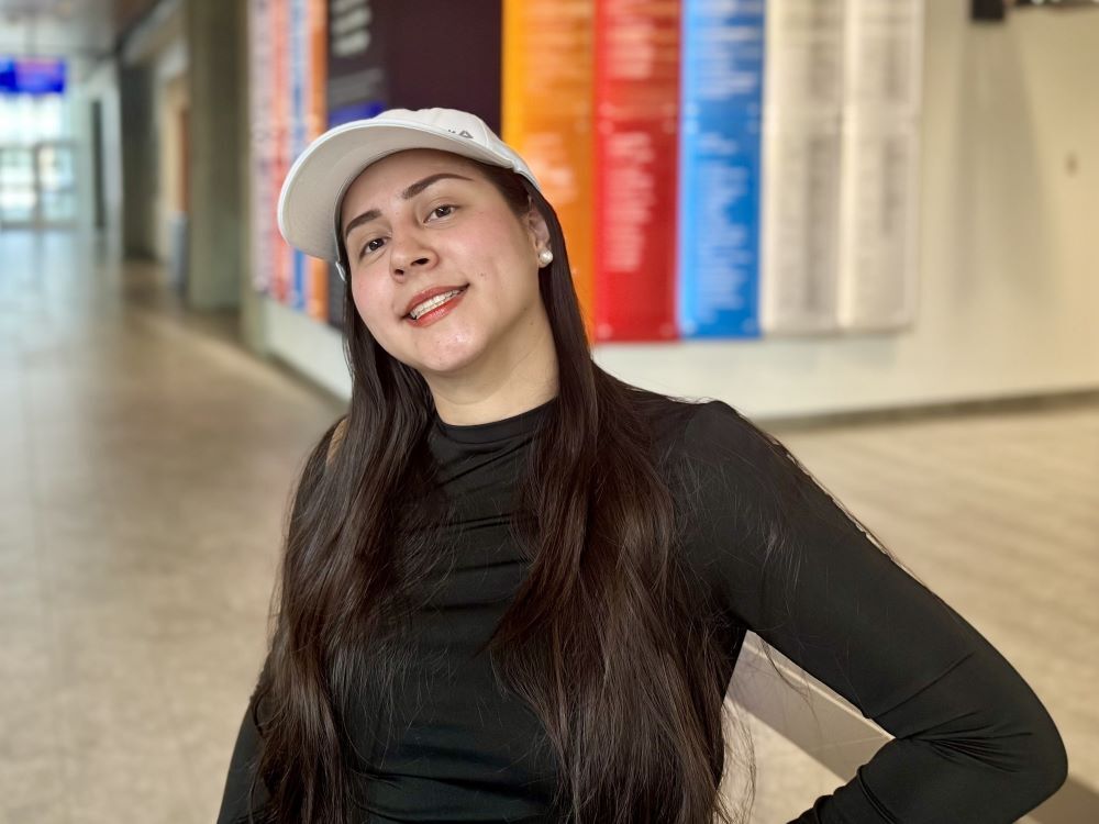 Samantha Mangabat is wearing a light ball cap and a long-sleeved black top. She has long dark hair and is smiling at the camera with her hand on her hip. She is standing in a hallway with a colourful vertical wall hanging in the background.