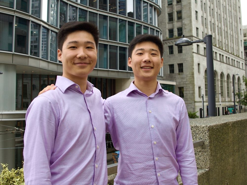 Twins Jerry and Jason Song have medium-light skin tone and dark hair. They are both wearing lilac-coloured button-up shirts. They are posing for a photo downtown near Waterfront Station in Vancouver.