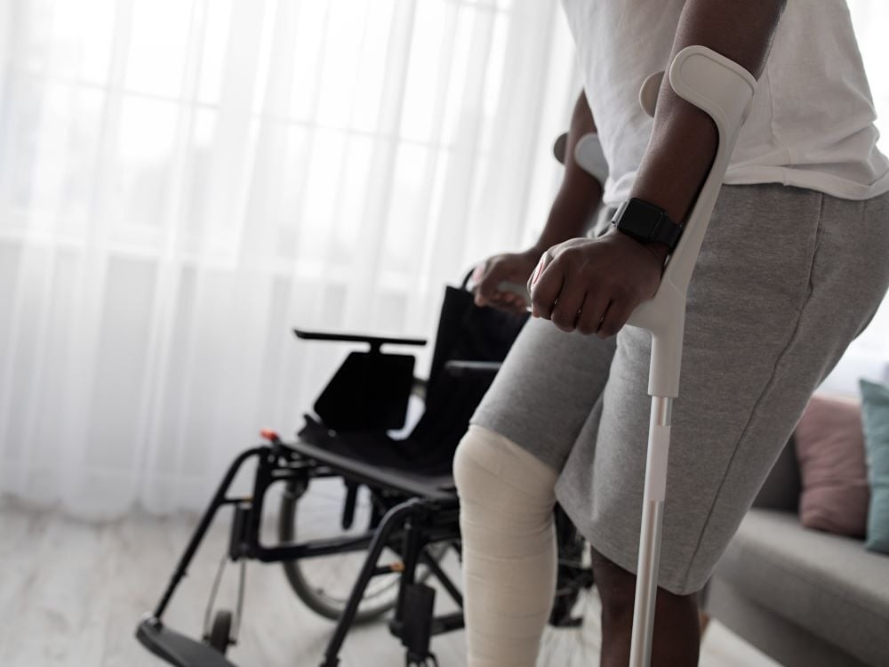 A dark-skinned man in sweatpants seen from the waist down is on crutches with a wheelchair in the background.