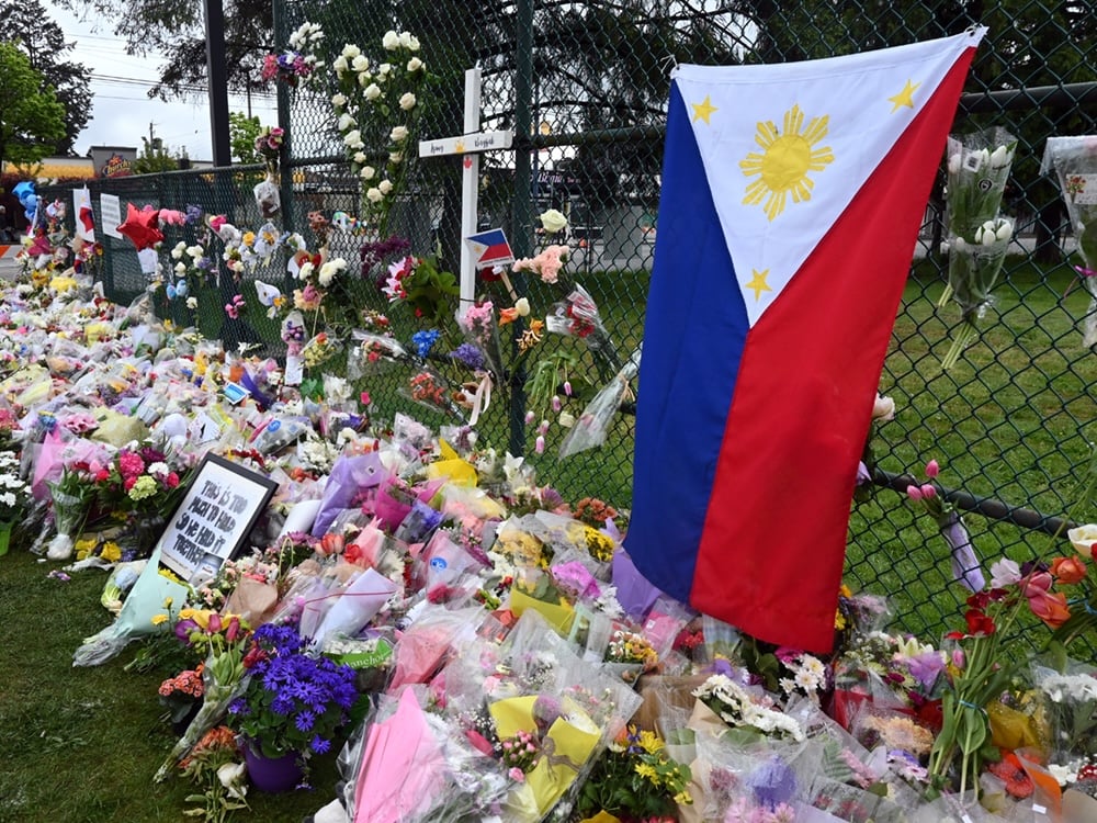 The flag of the Philippines is attached to a fence. It is surrounded by crosses, hearts and hundreds of bouquets of flowers.