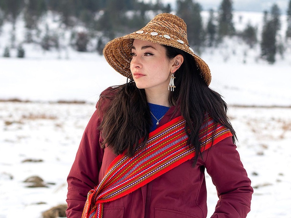A woman with light skin and long dark hair stands in a snowy field. She is wearing a woven Haida hat and a wine-coloured jacket.