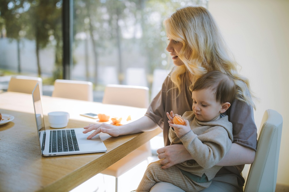 A blond-haired woman uses a computer while a toddler on her lap picks at a peeled orange.