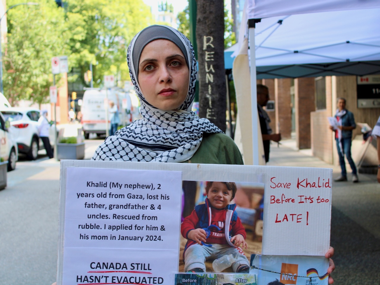 A woman with light skin tone wearing a head covering stands on a city sidewalk holding a cardboard poster bearing a young boy&rsquo;s photo and text including &lsquo;Save Khalid before it&rsquo;s too late!&rsquo;