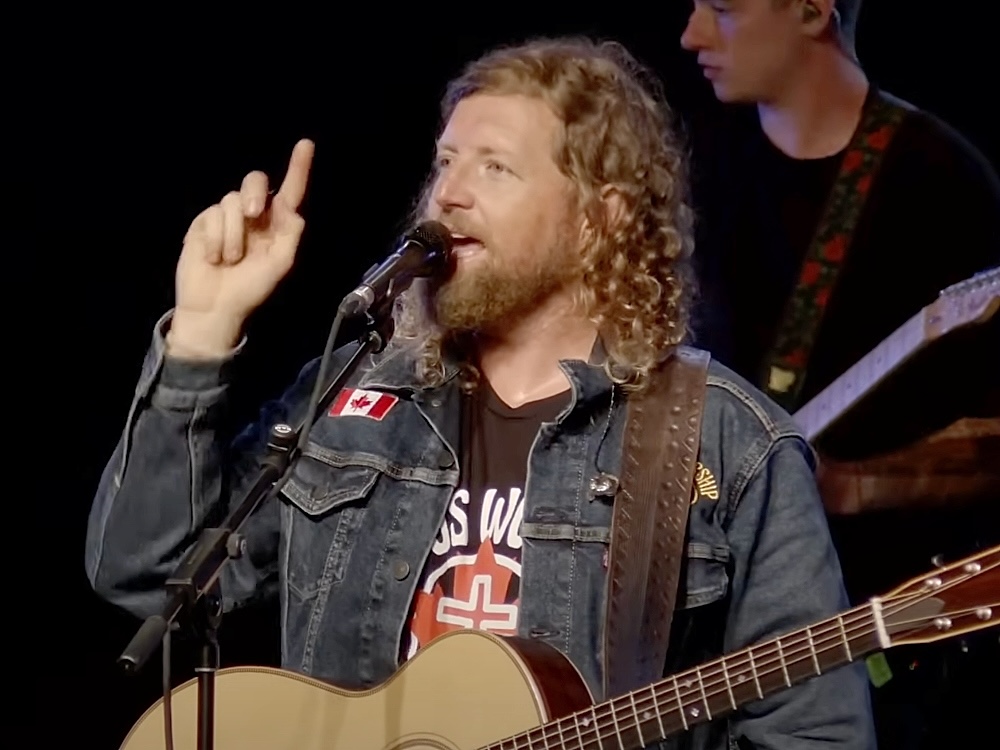 Sean Feucht has long curly blond hair and is holding an acoustic guitar, wearing a jean jacket with a Canadian flag. He is raising a finger while singing or talking into a microphone.