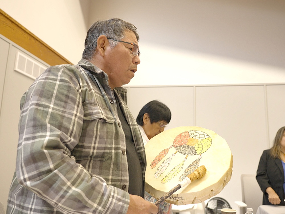 A man with medium skin tone, wearing a plaid shirt and glasses, plays a drum decorated with a medicine wheel dreamcatcher.